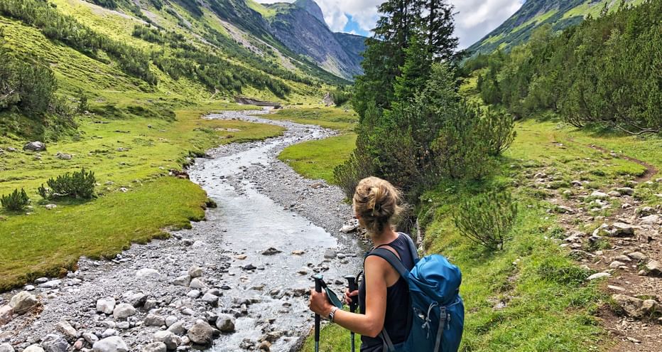 Female hiker with blue backpack and trekking poles on Lechweg, overlooking a green mountain valley with a winding stream.