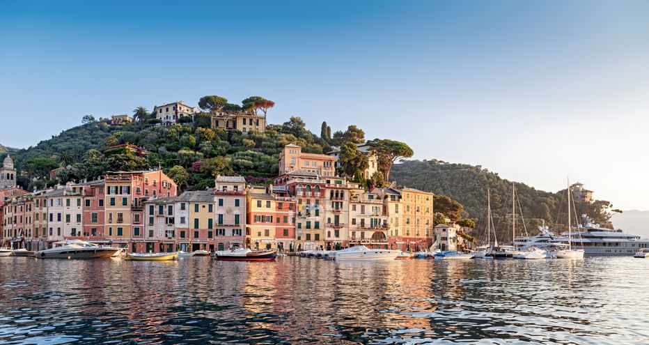 Hafen von Portofino mit bunten Pastellhäusern entlang der Uferpromenade, Boote und Yachten im ruhigen Wasser, Villen am Hang darüber.