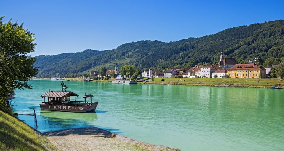 Traditional wooden ferry on turquoise Danube River near Engelhartszell with colorful buildings and forested hills in background under blue sky.