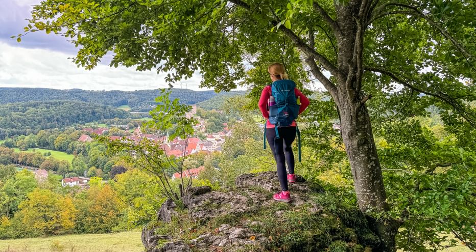 Wanderin mit Rucksack steht auf Felsvorsprung unter grünen Bäumen und blickt auf das Dorf Pappenheim im Altmühltal.