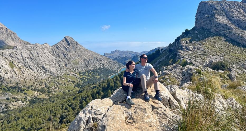 Two hikers sitting on rocky outcrop on GR221 trail in Mallorca, overlooking mountain valley with Puig Major peak and blue sky.