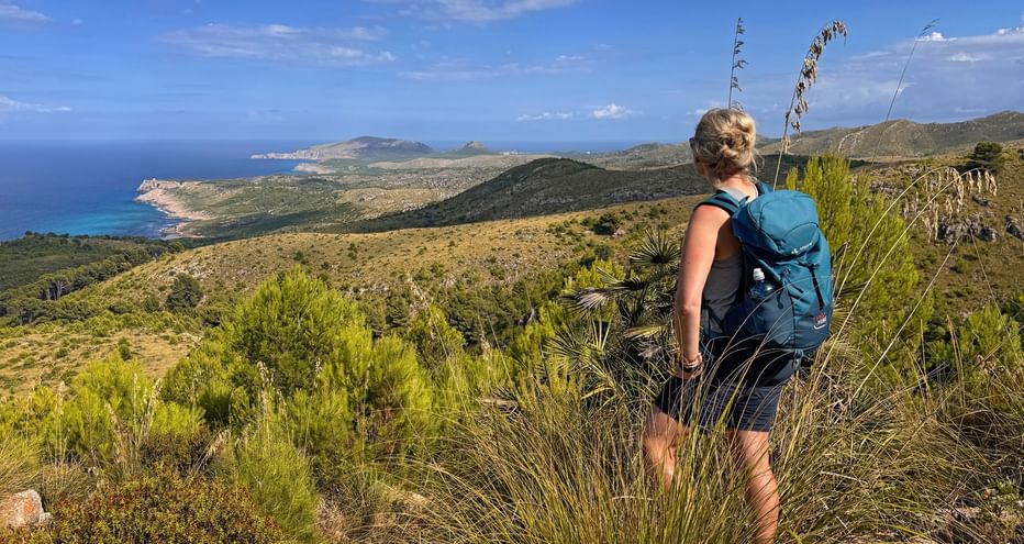 Wanderin mit blauem Rucksack auf Hügel mit Blick auf Mallorcas Ostküste mit türkisfarbenem Meer und bergiger Landschaft.