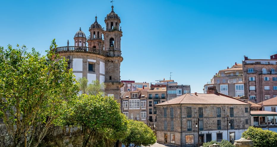 La Peregrina Kirche mit markanten Glockentürmen in Pontevedra, umgeben von Wohngebäuden und grünen Bäumen unter blauem Himmel.