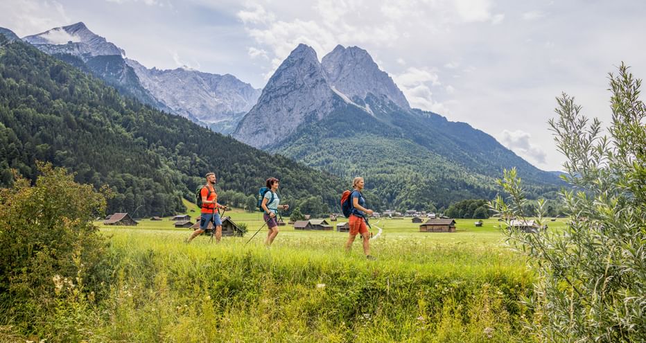 Drei Wanderer mit Rucksäcken gehen durch grüne Wiesen mit dramatischen Alpengipfeln im Hintergrund bei Garmisch-Partenkirchen.
