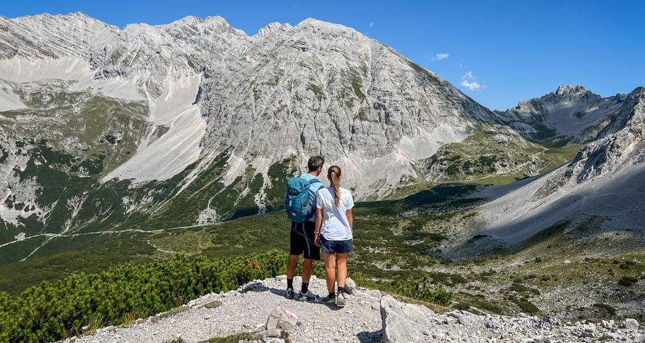 Two hikers standing on rocky viewpoint overlooking Karwendel mountain range with dramatic limestone peaks and green alpine valleys below.