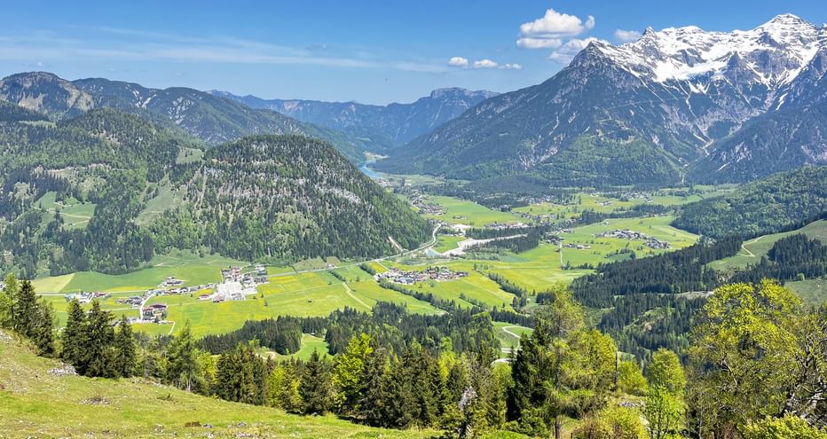 Alpine valley view from Buchensteinwand near Kitzbühel with green meadows, forested hills, small villages, and snow-capped mountains under blue sky.