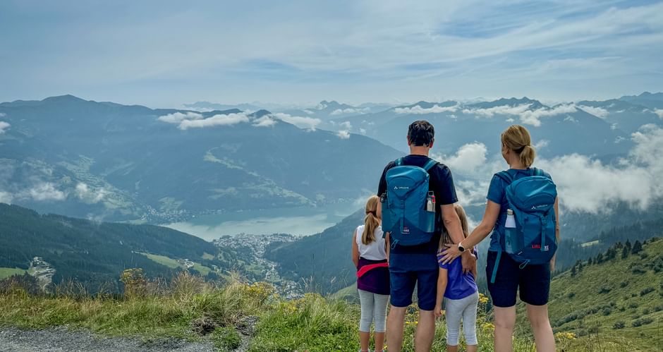 Family of four with backpacks viewing alpine panorama in Pinzgau. Mountain lake and peaks visible below through clouds.