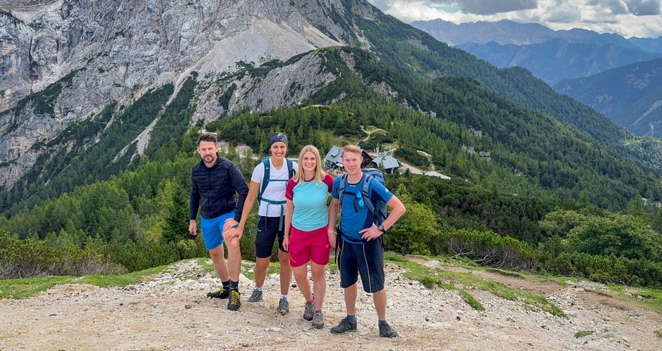 Vier Wanderer mit Rucksäcken auf dem Vršič-Pass-Gipfel mit bewaldeten Bergen und felsigen Gipfeln der Julischen Alpen im Hintergrund.