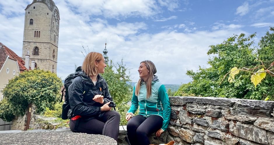 Two women hikers with a dog resting on a stone wall in Stein an der Donau, Wachau. Historic church tower and vineyard landscape visible.