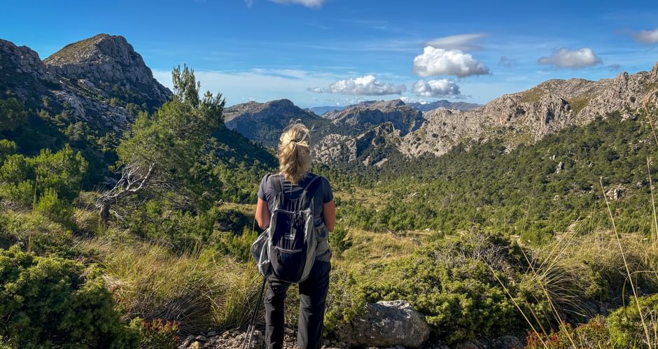 Hiker with backpack standing on rocky trail overlooking Tramuntana mountain range with green valleys and blue sky with white clouds.