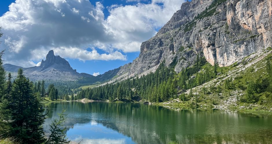 Lago Federa spiegelt dramatische Dolomitengipfel und Wolken. Tannenwälder umgeben den grünen Alpensee mit steilen Felsklippen rechts.