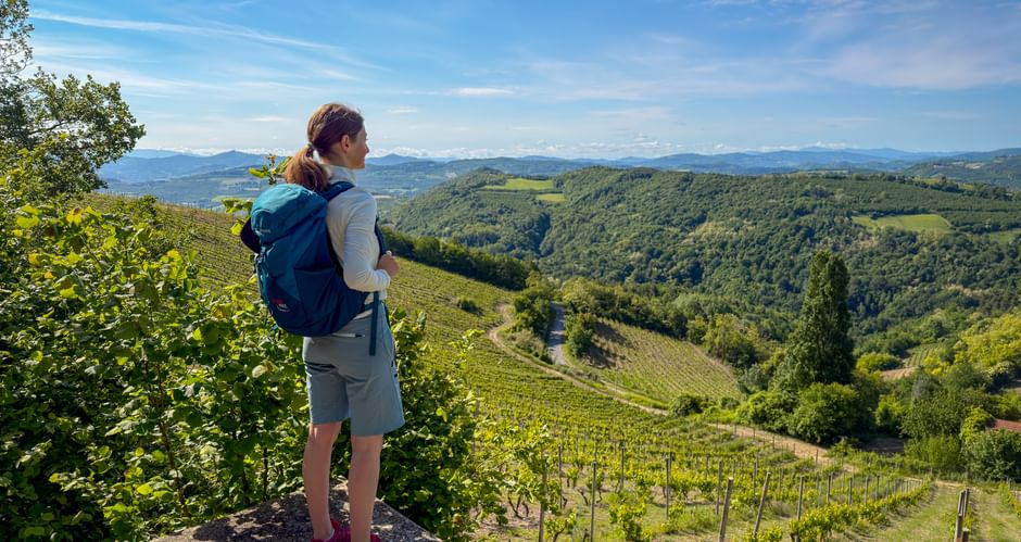 Female hiker with blue backpack standing on rock near Benevello, overlooking rolling hills with vineyards in Piedmont under blue sky.