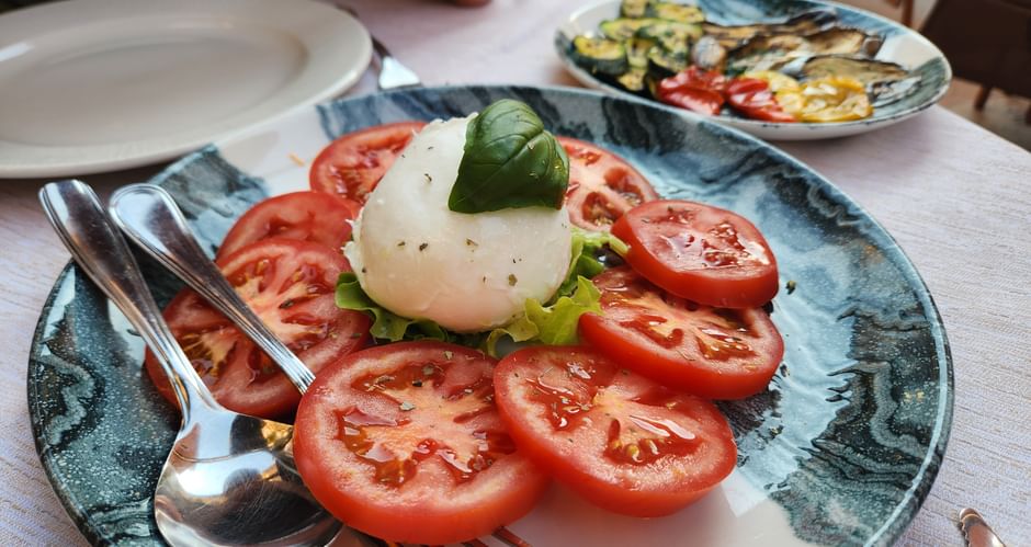 Caprese salad with sliced tomatoes, mozzarella, and basil on a blue plate. Grilled vegetables visible on a second plate in the background.