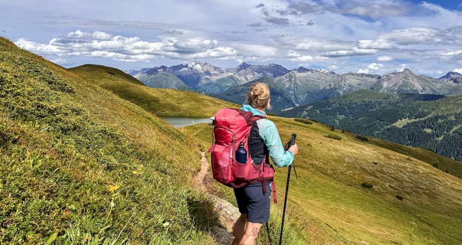 Wanderer mit rotem Rucksack und Wanderstöcken auf Bergweg bei Obernberg mit Blick auf Alpengipfel unter blauem Himmel mit Wolken.