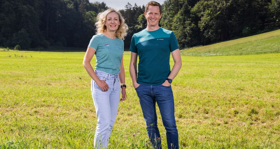 Two people Verena Sonnenberg und Thomas Schmid standing in a green meadow with forest in the background. Both wear blue outdoor shirts and smile at the camera.