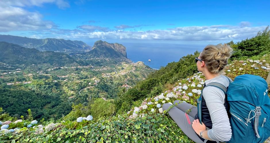 Female hiker with blue backpack sitting on hillside overlooking Madeira's north coast with dramatic cliffs, ocean, and white flowers in foreground.