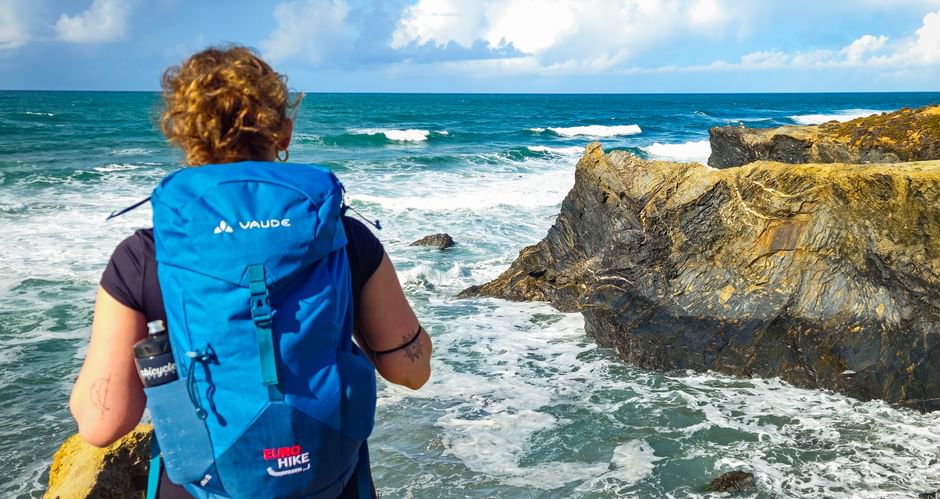Wanderin mit blauem Rucksack betrachtet felsige Küste an der Praia das Furnas an der Rota Vicentina. Wellen brechen an goldenen Felsen unter blauem Himmel.