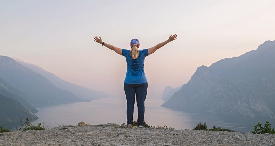 Woman with raised arms standing on rocky summit at Monte Brione overlooking Lake Garda at sunset, mountains visible on both sides.