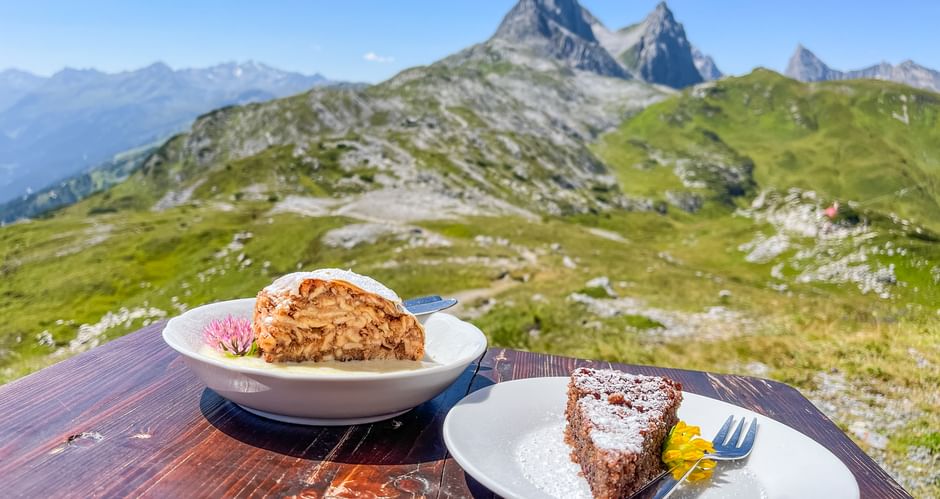Two dessert slices on white plates at a mountain hut table, with green alpine meadows and rocky peaks of the Allgäu-Lechtal Alps behind.