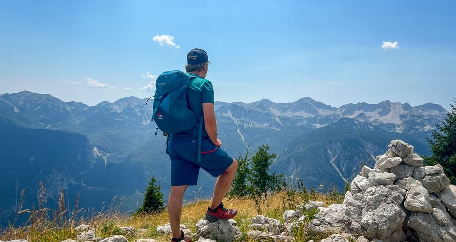 Hiker with backpack standing on rocky summit in the Julian Alps, overlooking mountain ranges under blue sky with stone cairn nearby.
