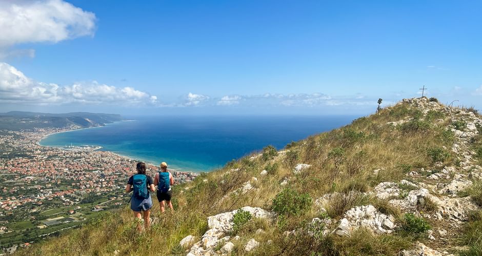 Two hikers walking uphill on Monte Croce with coastal views of Piedmont-Liguria. A summit cross is visible on the rocky peak under blue sky.