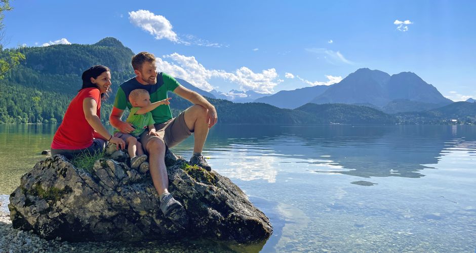 Family of three taking a break from hiking at the lake, sitting on a rock with a view of the Dachstein in the background