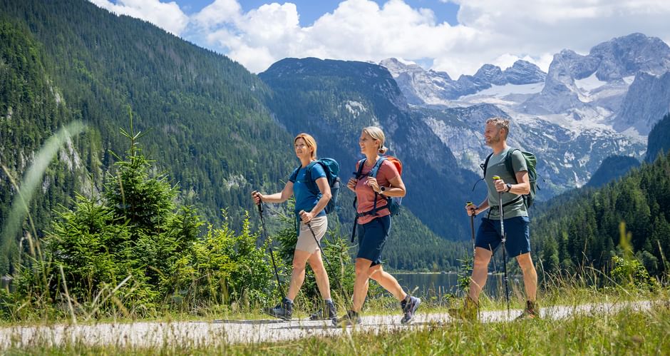 Drei Wanderer mit Rucksäcken auf einem Weg am Gosausee mit dem schneebedeckten Dachstein-Gebirge und bewaldeten Hängen im Hintergrund.