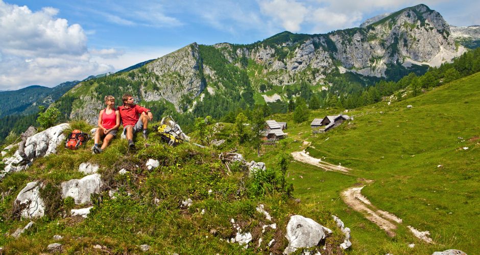 Wanderpaar bei der Rast auf der Krstenica Alm mit Hütten und Bergen im Hintergrund