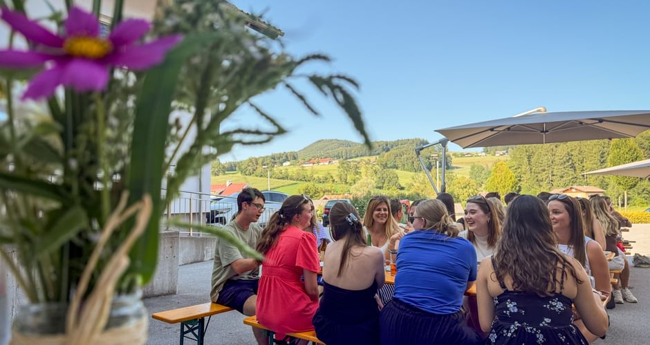Mitarbeiter sitzen an Biergarnituren bei einem Sommerfest im Freien. Rosa Blumen im Vordergrund, Hügel und blauer Himmel im Hintergrund.