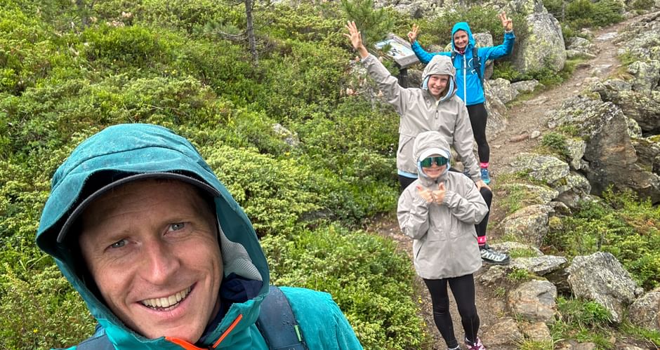 Smiling man taking selfie with family on rocky mountain trail in rain. Three people behind wear rain jackets, surrounded by green vegetation.