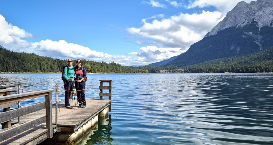 Zwei Wanderer stehen auf einem Holzsteg am Eibsee mit klarem türkisfarbenem Wasser, bewaldeten Ufern und der Zugspitze im Hintergrund.