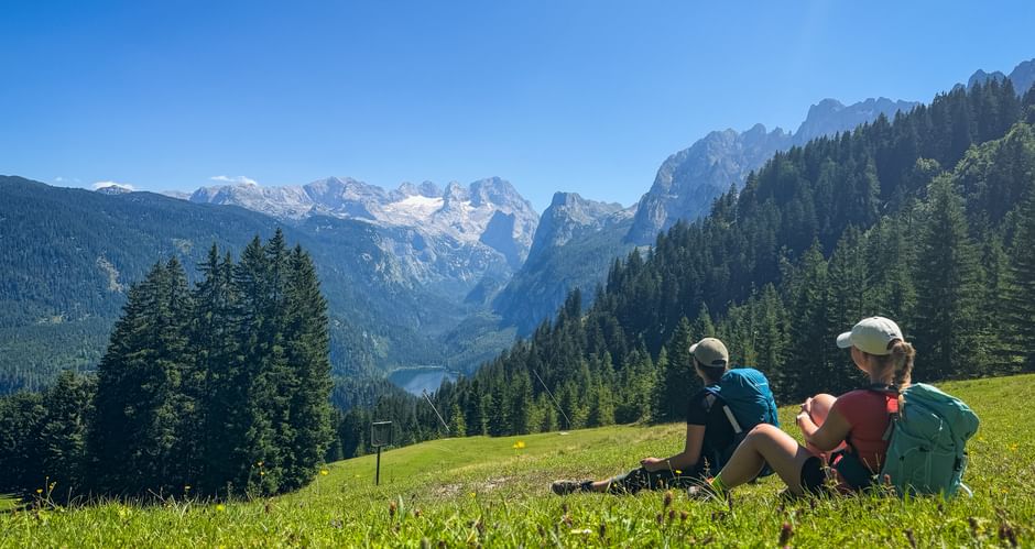 Two hikers with backpacks sitting on a green meadow in Gosau, viewing the Dachstein glacier and mountain peaks under a clear blue sky.