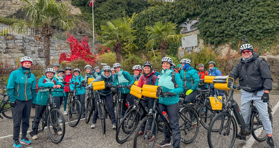 Large group of cyclists in turquoise jackets with helmets and bikes with yellow panniers, posing in front of stone wall with palm trees.