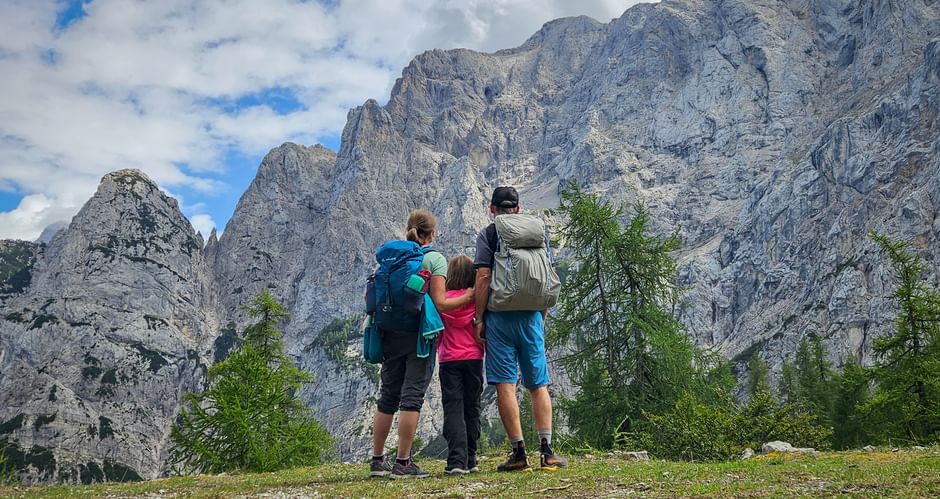 Family of three with backpacks viewing dramatic rocky peaks of the Julian Alps. Green meadow and trees in foreground, cloudy blue sky above.