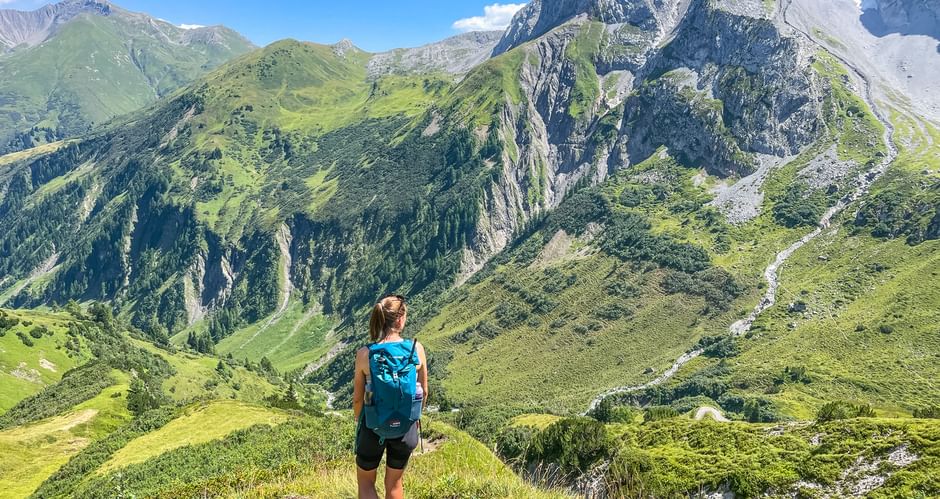 Wanderer mit blauem Rucksack auf Bergpfad mit Blick auf grüne Täler und felsige Gipfel in den Allgäuer und Lechtaler Alpen.