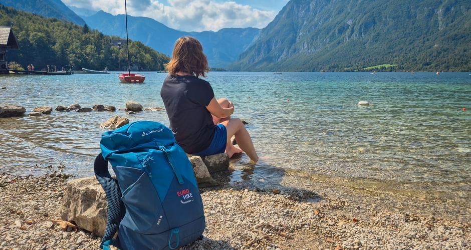 Wanderer mit blauem Rucksack sitzt auf Felsen am Ufer des Bohinjer Sees. Türkisfarbenes Wasser, Segelboot und Julische Alpen unter blauem Himmel.