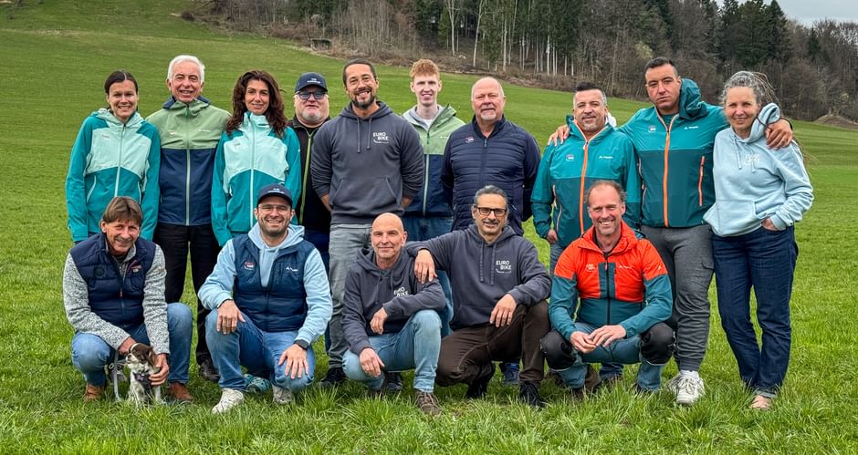 Group photo of 16 people in outdoor clothing on a green meadow. Rolling hills and forest visible in the background under cloudy sky.