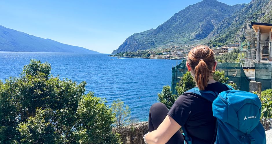 Wanderin mit blauem Rucksack blickt auf den Gardasee und Limone sul Garda, mit Bergen und blauem Wasser unter klarem Himmel.