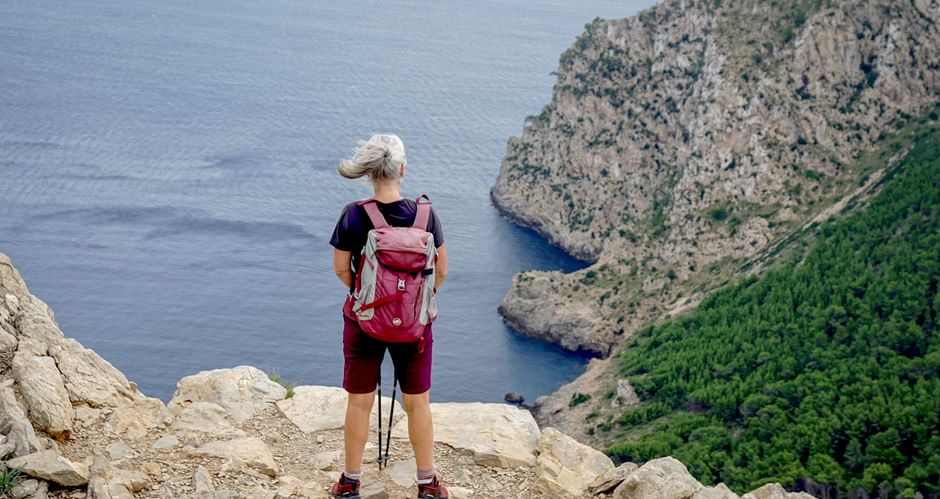 Female hiker with backpack and trekking poles standing on rocky cliff overlooking the Mediterranean Sea near Alcudia, Mallorca.