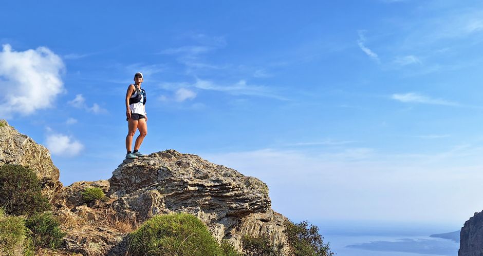 Woman stands in sportswear on a rock high above the sea, blue sky, sunshine