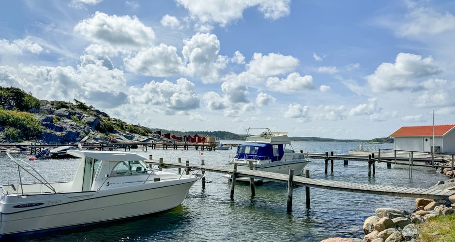 Weiße Motorboote an Holzstegen im Hafen von Särö. Felsige Küste im Vordergrund, Gebäude mit rotem Dach sichtbar, blauer Himmel mit weißen Wolken.