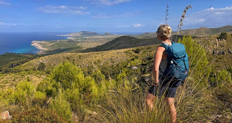 Wanderin mit blauem Rucksack auf Hügel mit Blick auf Mallorcas Ostküste mit türkisfarbenem Meer und bergiger Landschaft.