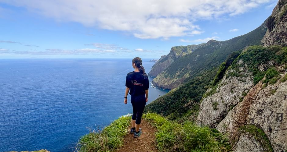 Female hiker walking on Vereda do Larano coastal trail in Madeira with steep cliffs, green vegetation, and deep blue Atlantic Ocean.