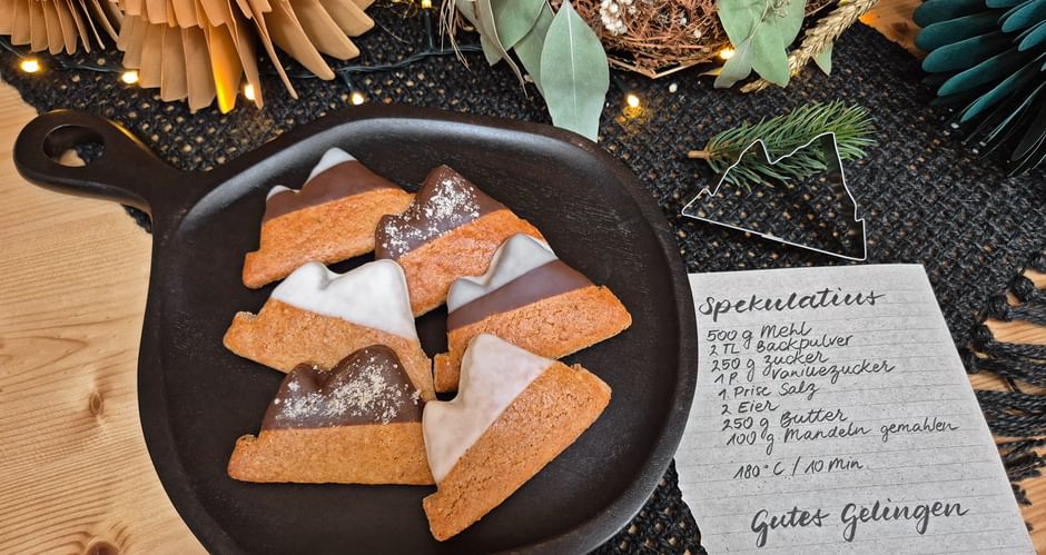 Cast iron pan with glazed speculatius cookies beside a handwritten recipe card, surrounded by festive decorations including paper fans and greenery.
