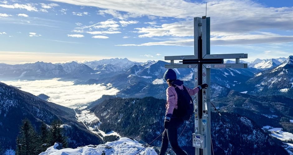 Wanderer in lila Jacke steht am Gipfelkreuz auf schneebedecktem Gipfel mit Blick auf verschneite Alpentäler und Bergketten unter blauem Himmel.