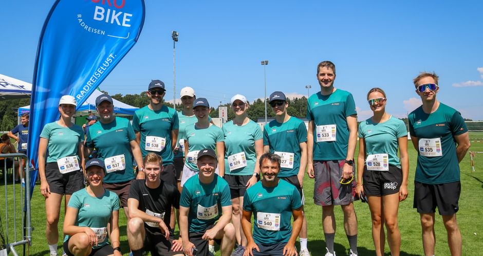 Group of runners in turquoise shirts with race bibs posing on grass field. Eurobike flag visible on left under clear blue sky.