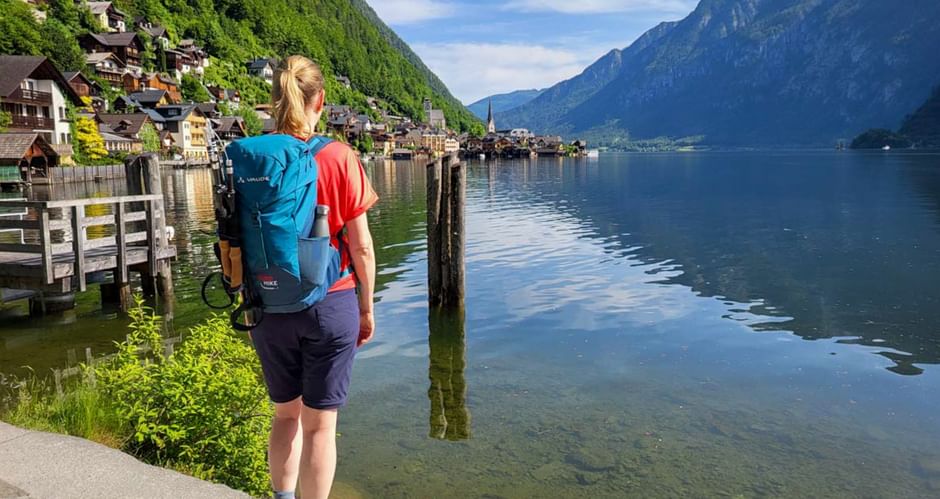 Female hiker with blue backpack viewing Hallstatt village across calm lake waters, surrounded by steep forested mountains under blue sky.